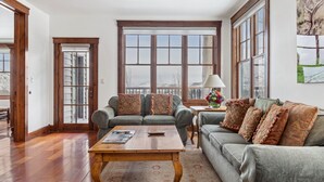 A living room with a wooden coffee table, two grey sofas with patterned cushions, a side table with a lamp, and large windows with a view of a snowy landscape.