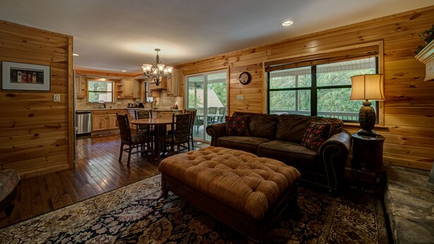 A cozy cabin interior with wooden walls, a dining area, a living room with a sofa and ottoman, and large windows offering a view of the outdoors. - LAKEFRONT, PONTOON BOAT, HOT TUB, FIREPIT, Fireplace . Wide lake Mountain View  (Blairsville)