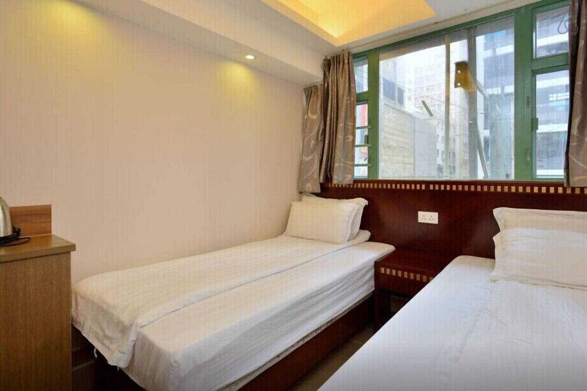 A hotel room with two single beds, a wooden nightstand, a window with curtains, and a view of buildings outside.