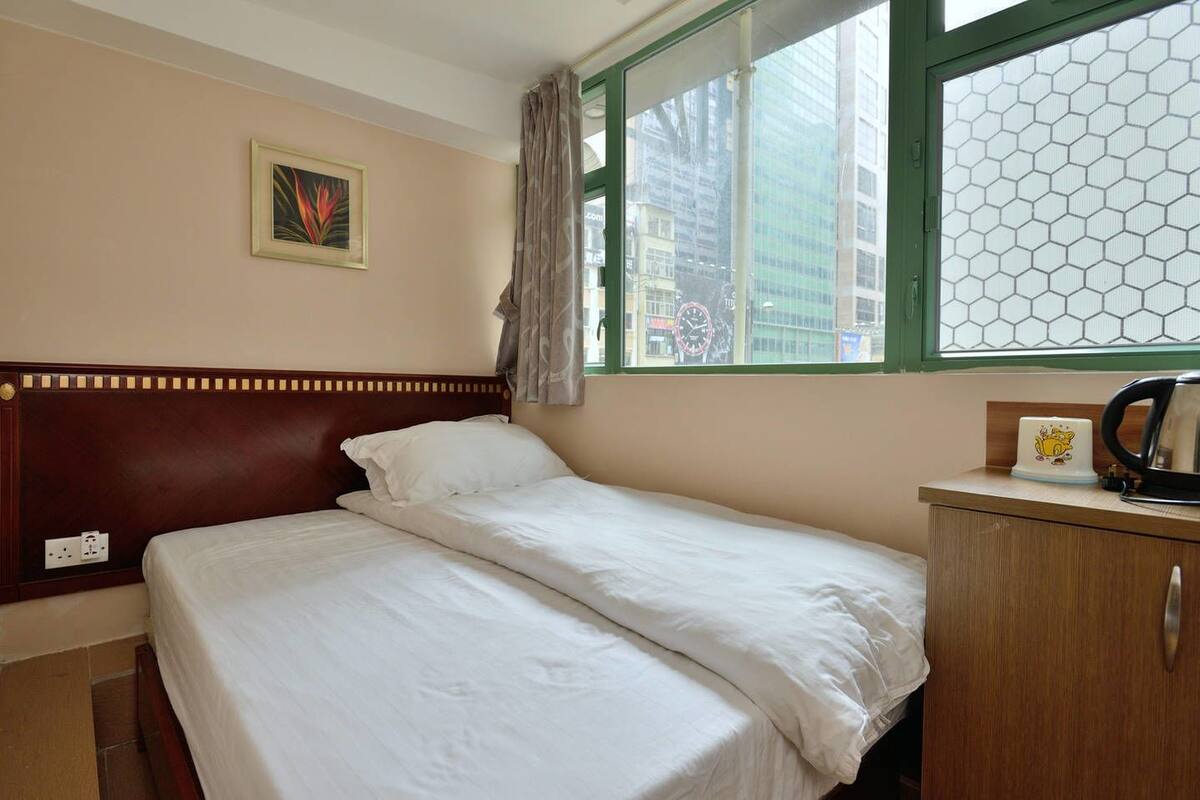 A single bed with white bedding, a wooden headboard, a small bedside table with a kettle and a clock, a window with a green frame, and a framed picture on the wall.