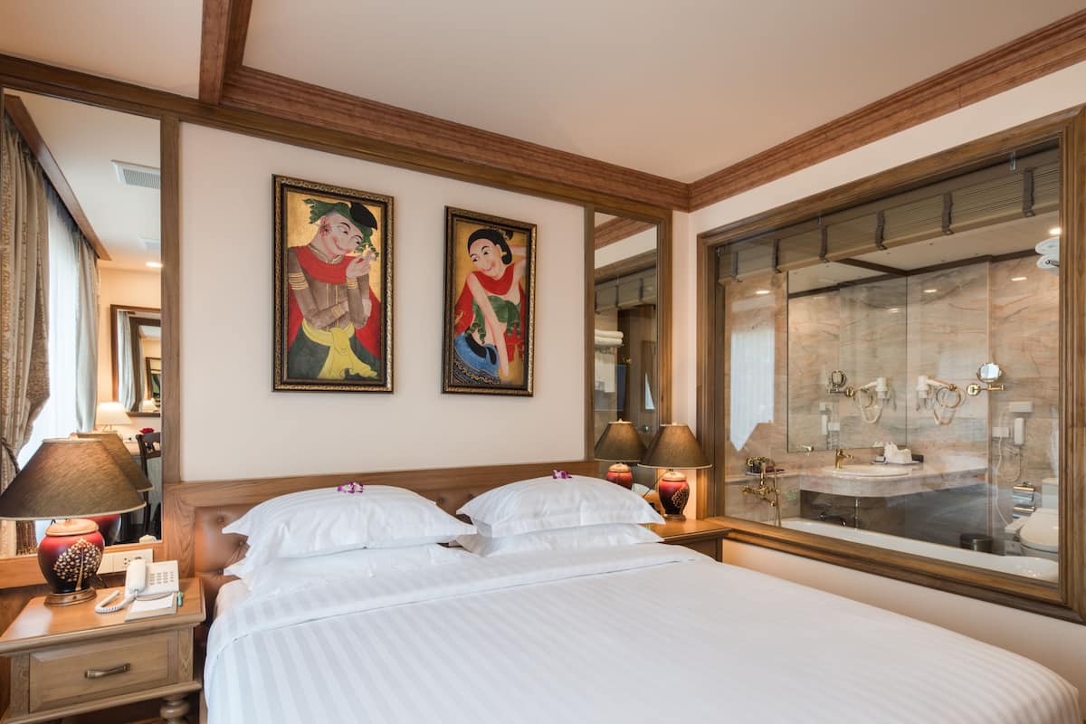 A hotel room with a large bed, a nightstand with a lamp, a telephone, and a mirror. There are two paintings on the wall and a bathroom visible through a glass door.