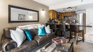 Een woonkamer met een leren bank, een salontafel en een keukenhoek met houten kasten en een roestvrijstalen koelkast.