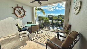 A balcony with a table and chairs, a ceiling fan, a ship's wheel decoration, and a view of a pool and palm trees. - Another Day in Paradise           Best Waterfront and Poolside  Views. (Key Largo)