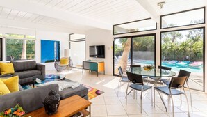 A modern living room with a sofa, armchair, and dining table, large windows, and a pool view. - Desert Modern by Jack Meiselman with Pool/Spa (Palm Springs)