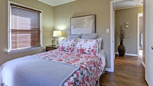 A bedroom with a bed, a nightstand, a lamp, a painting, and a vase with branches.