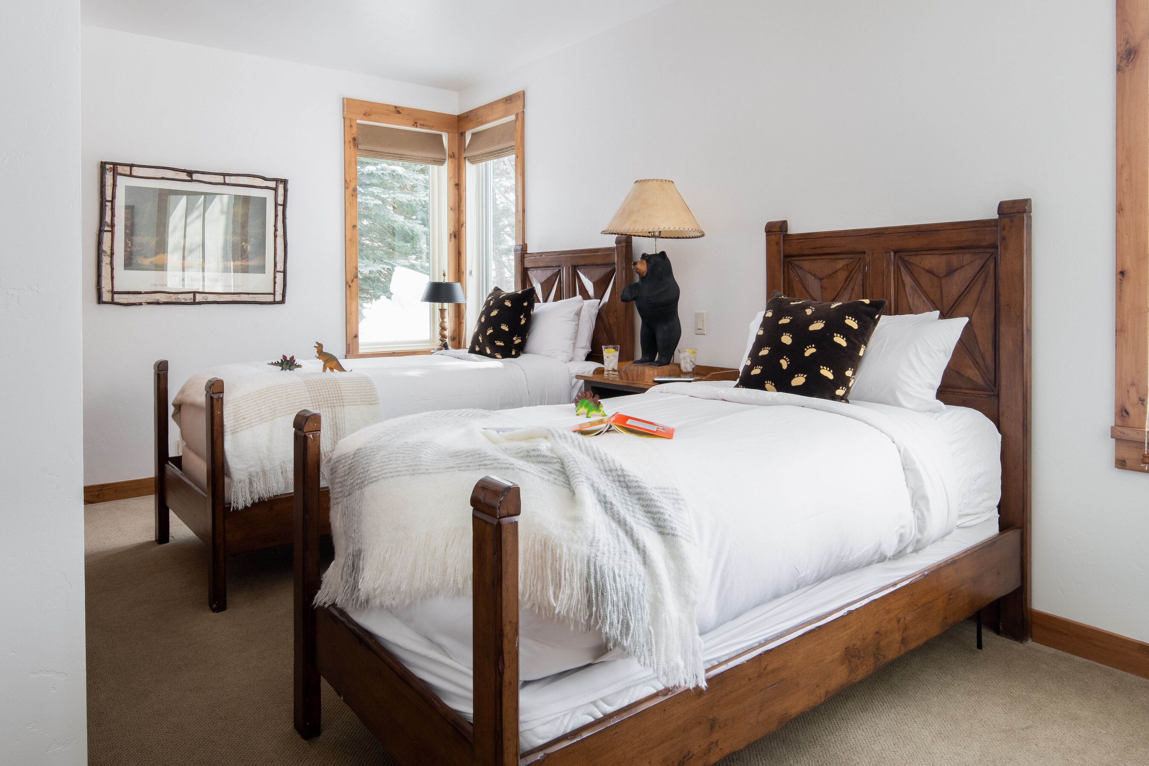 A bedroom with two single beds, wooden headboards, a framed picture on the wall, and a window with a view of trees.
