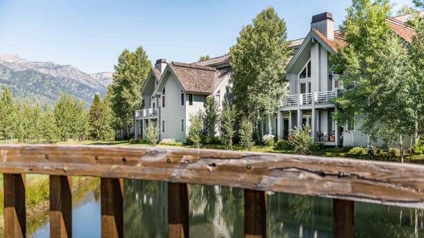 A large, modern house with a prominent balcony, situated next to a calm lake with mountains in the background. - Outpost: Granite Canyon at Teton Pines (Wilson)