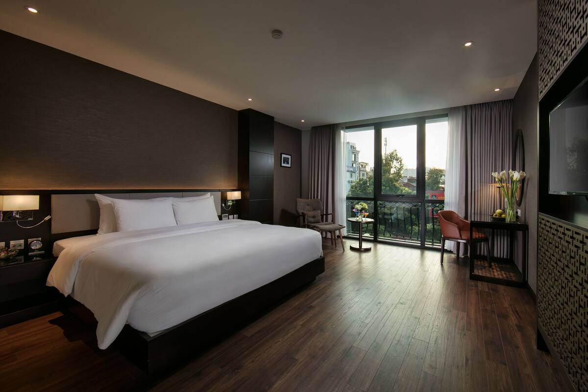 A modern hotel room with a large bed, a small table with flowers, a chair, and a balcony with a view.