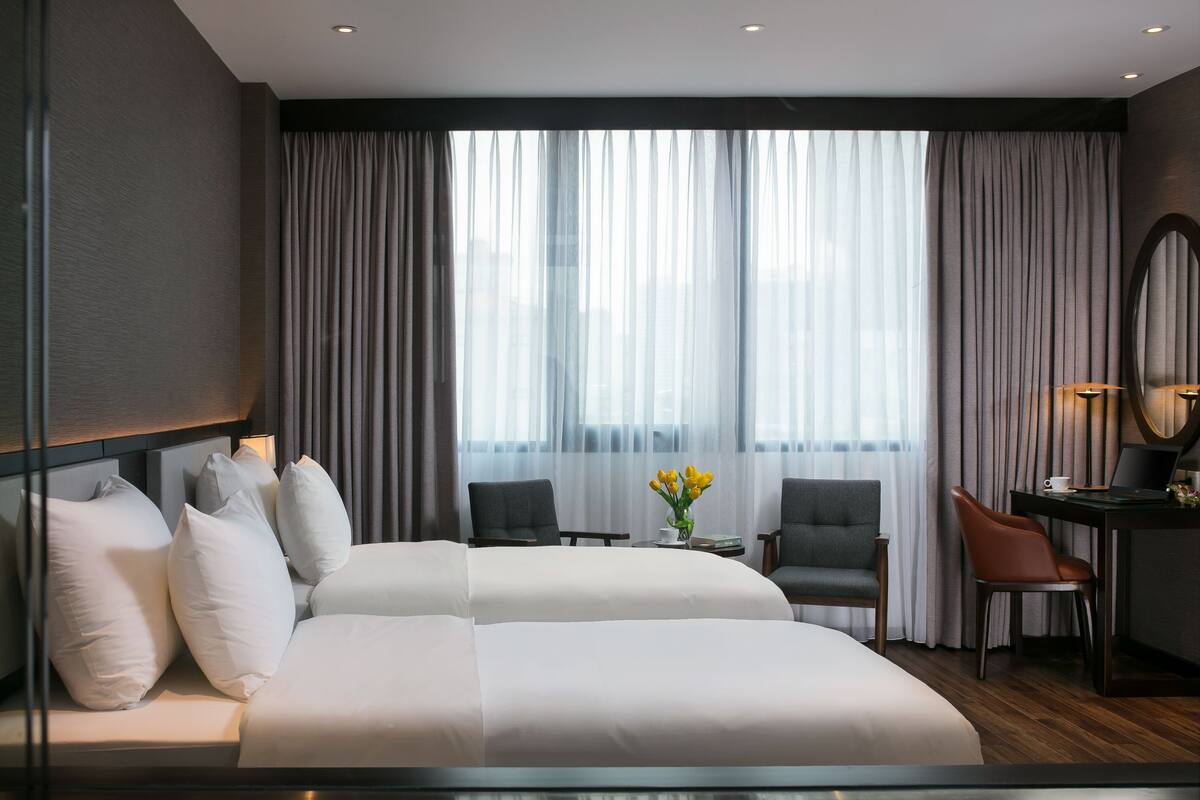 A modern hotel room with a large bed, two chairs, a desk with a laptop, and a view of the city through sheer curtains.