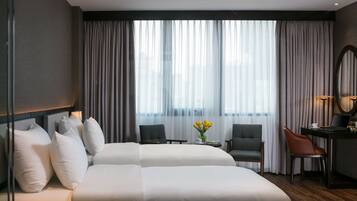Habitación de hotel moderna con una cama grande, dos sillas, un escritorio con una computadora portátil y vistas a la ciudad a través de cortinas translúcidas.