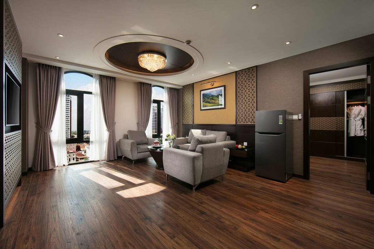 A modern hotel room with a wooden floor, a sofa, a dining area, and a view of the cityscape.
