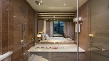 Un baño moderno con ducha de vidrio, una bañera con pétalos de rosa y vistas a un dormitorio con cama y mesita de noche.