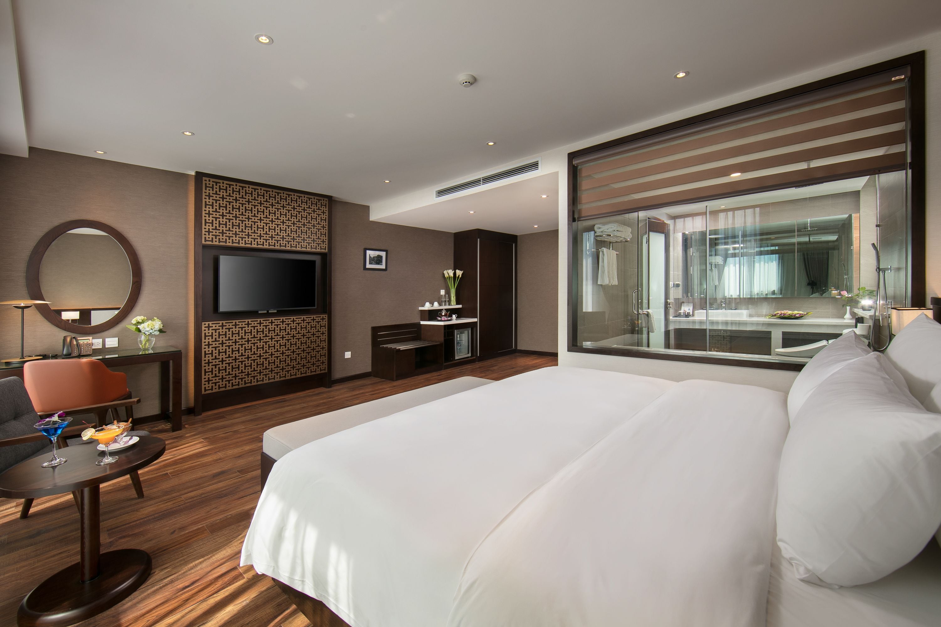 A modern hotel room with a large bed, a TV, a desk with a chair, and a view of the bathroom through a glass partition.