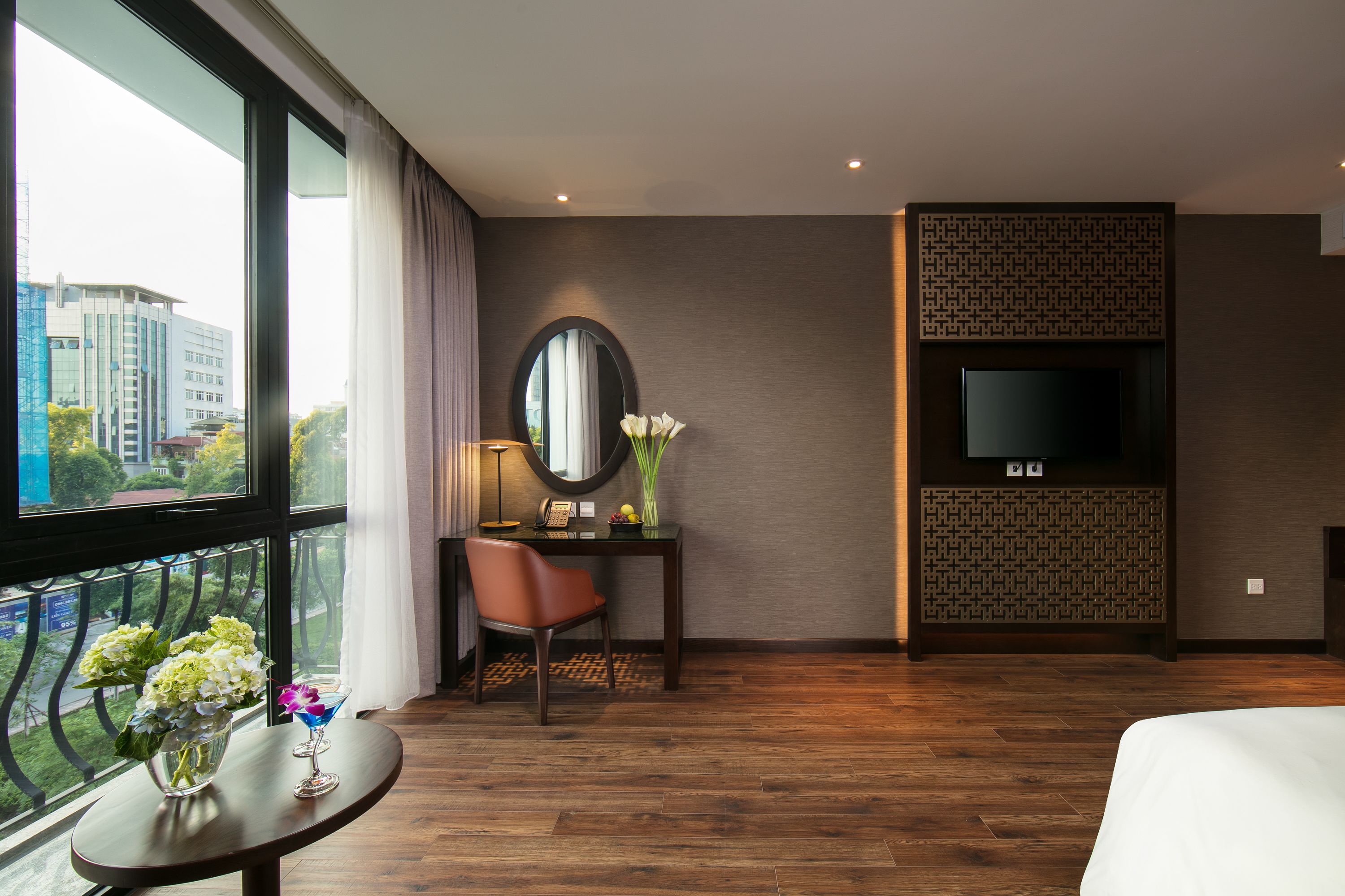 A hotel room with a large window, a desk with a chair, a TV mounted on a wall, and a bed.