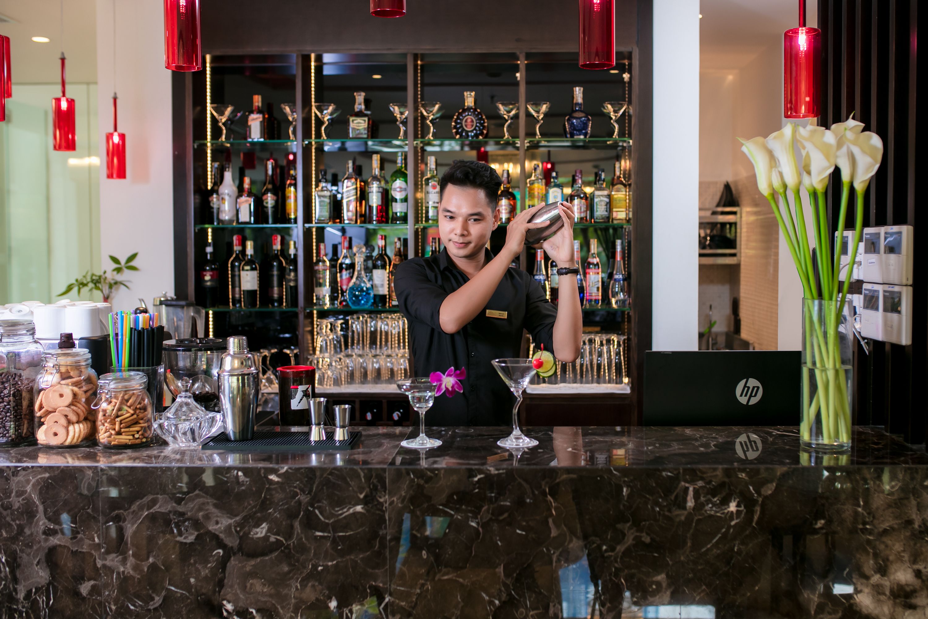 A bar area with a bartender preparing a drink, shelves with liquor bottles, and a vase with white flowers.