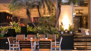 Un vestíbulo de hotel con mostrador de recepción, zona de estar con sillas y mesas de madera, y varios maceteros con plantas.