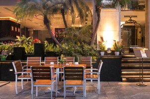 A hotel lobby with a reception desk, seating area with wooden chairs and tables, and a variety of potted plants.