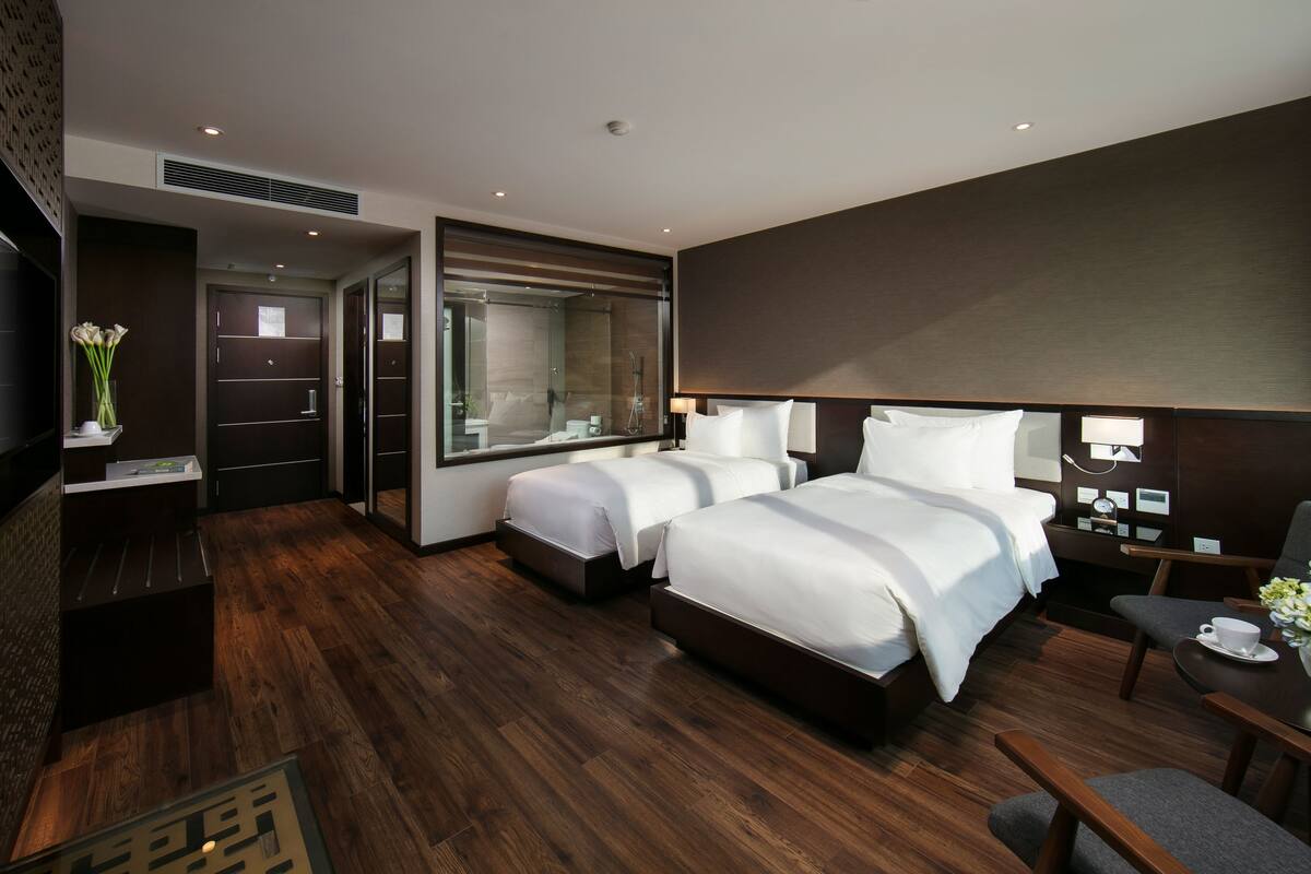 A modern hotel room with two beds, a desk, a chair, a TV, and a bathroom visible through a glass door.