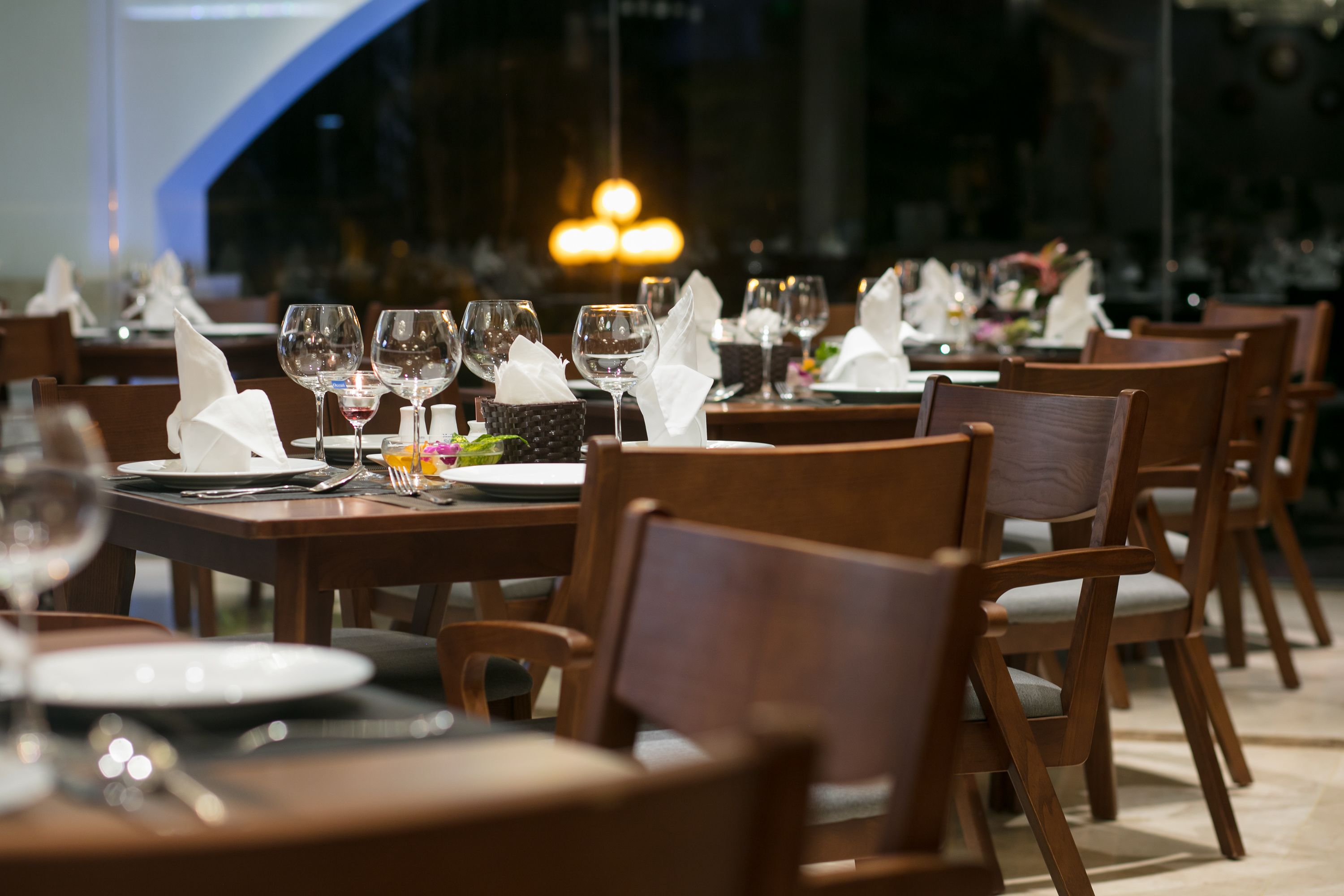 A restaurant with wooden tables and chairs, each set with plates, cutlery, and wine glasses.