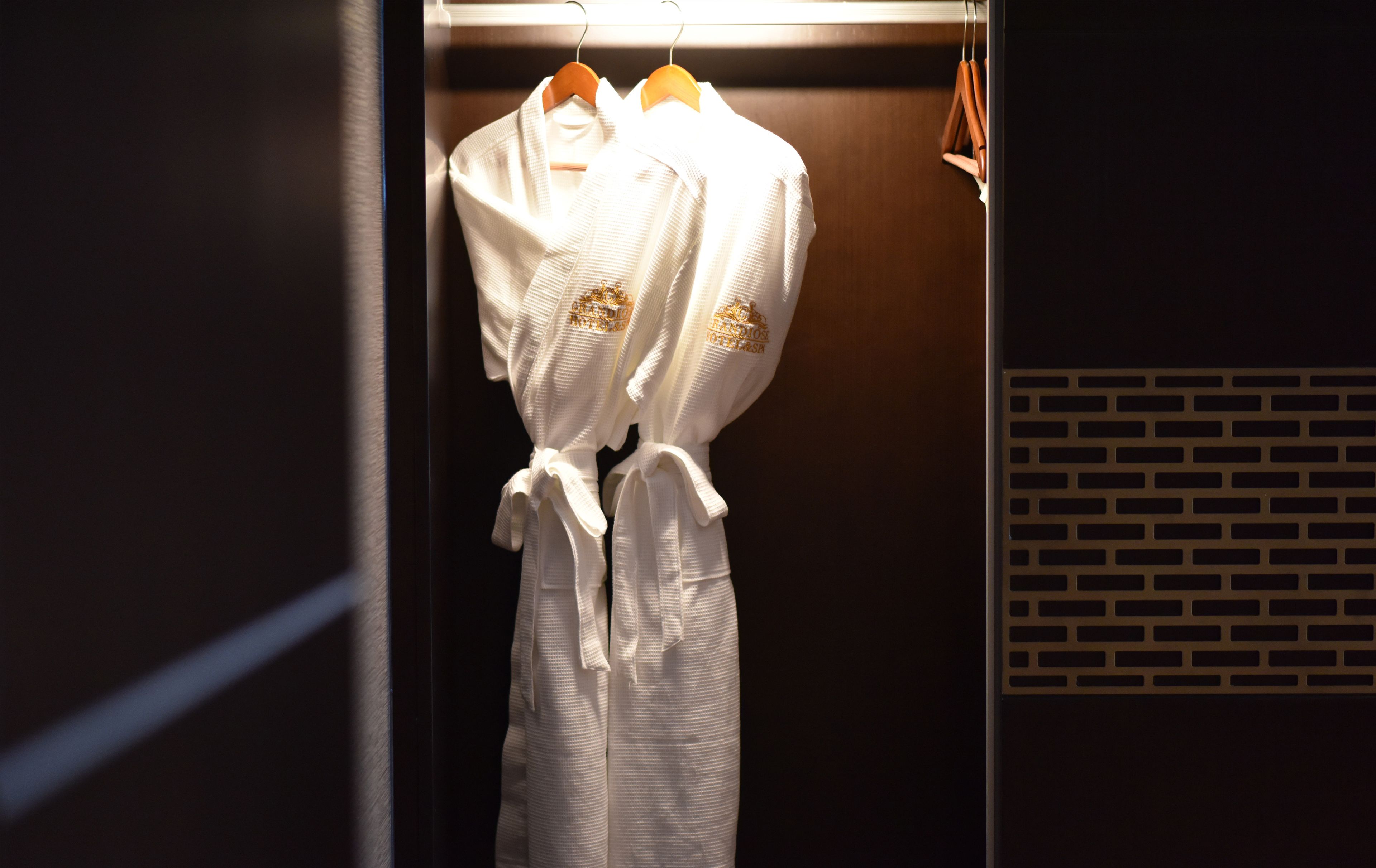 Une robe blanche est suspendue à un cintre en bois dans une pièce faiblement éclairée, dont un mur est en briques et qui est équipée d’une grille de ventilation en métal.