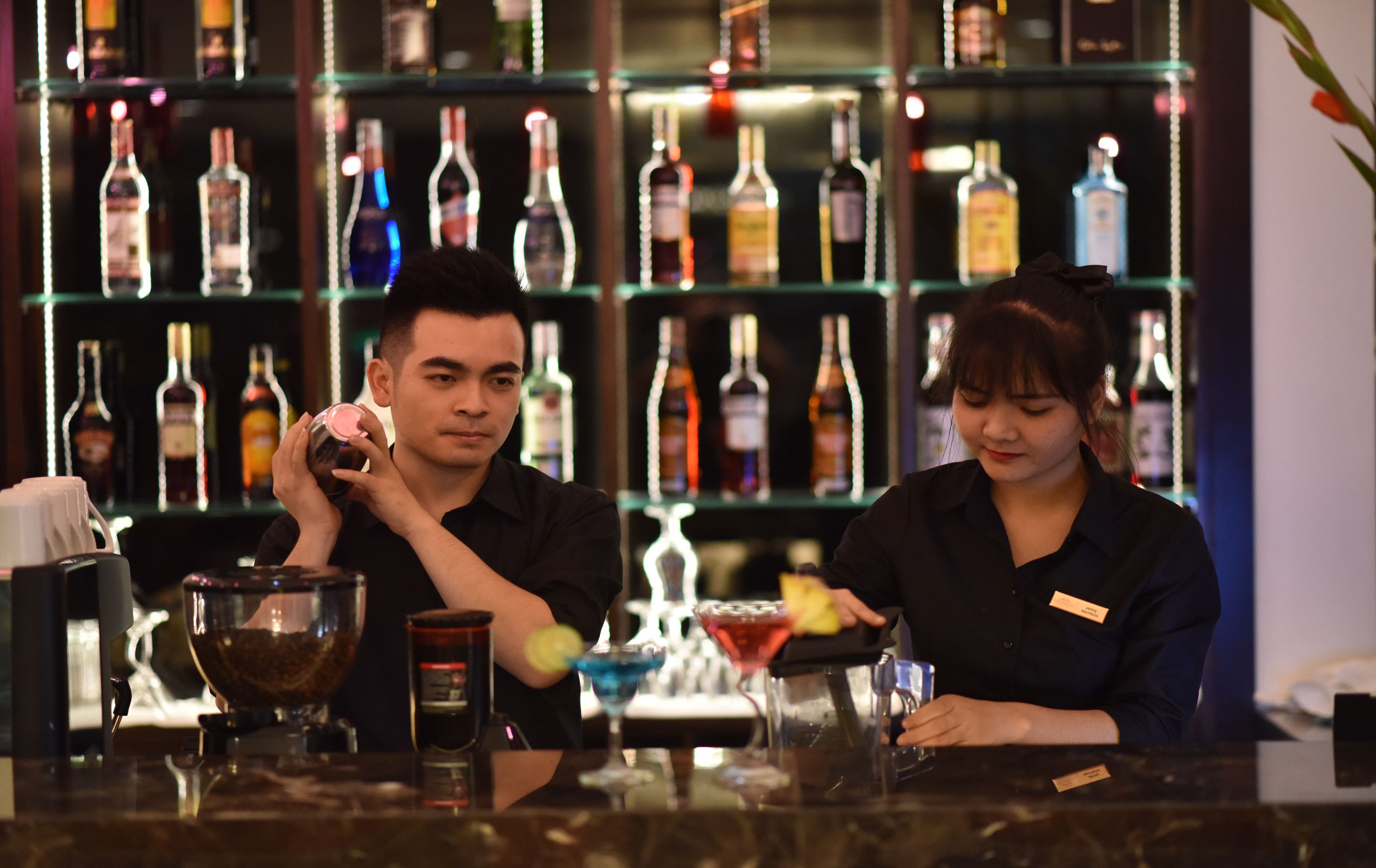 Eine Bar mit einem Barkeeper, der ein Getränk zubereitet. Im Hintergrund sind Regale mit verschiedenen Flaschen zu sehen.