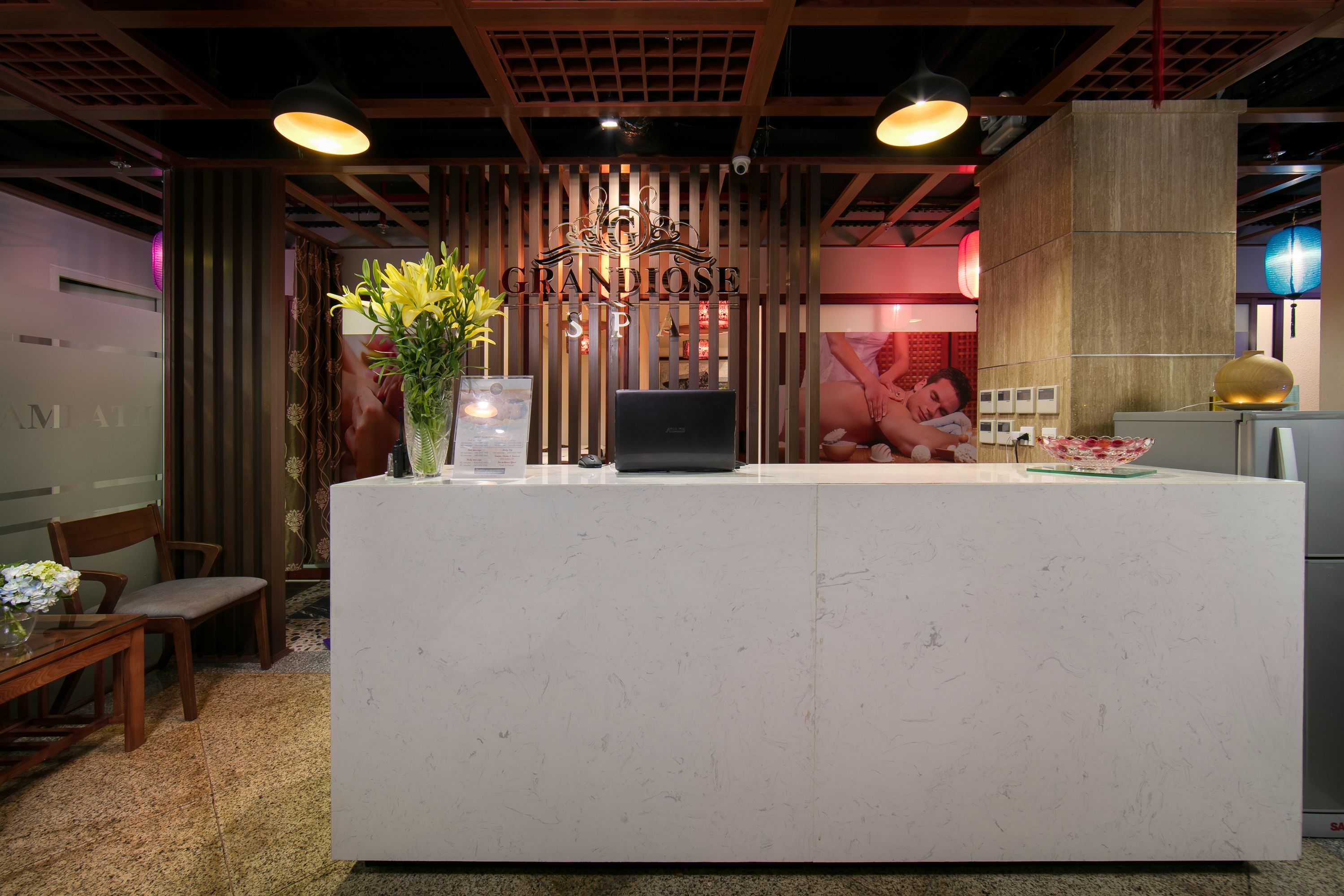 A reception desk with a laptop, a vase with flowers, and a wooden partition with the name "GRANDIOSE SPA".