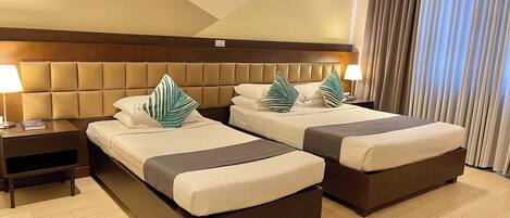 A hotel room with two beds, a wooden headboard, a nightstand with a lamp, and a large window with curtains.