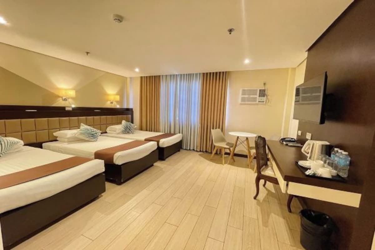 Habitación de hotel con tres camas, un rincón de comedor pequeño y un televisor montado en la pared.