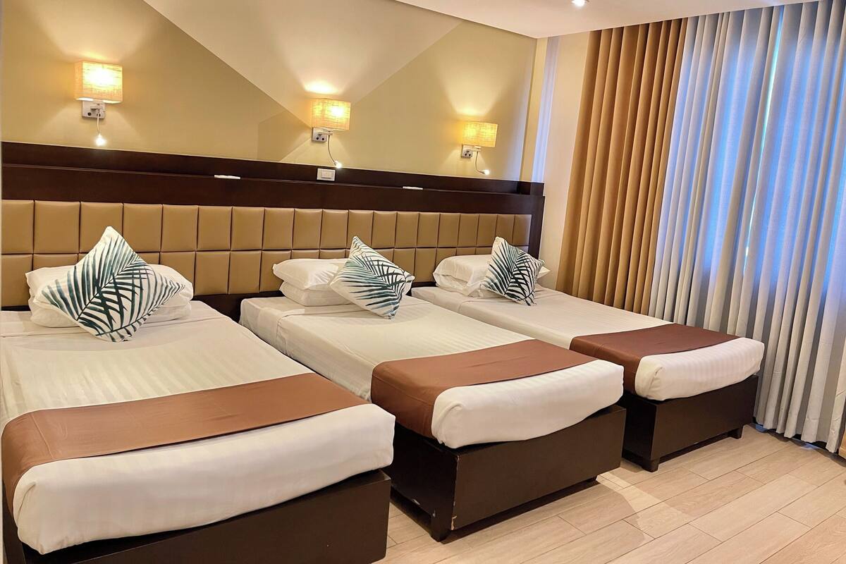 A hotel room with four beds, each with a brown headboard and white bedding. There are wall-mounted lights and a large mirror above the beds.