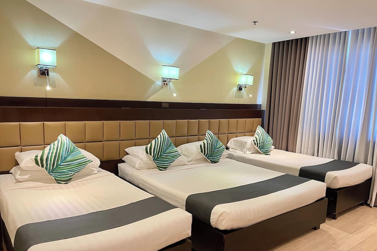 A hotel room with three beds, each with a dark headboard and white bedding. There are two wall-mounted lights above each bed, and a large window with curtains to the right.