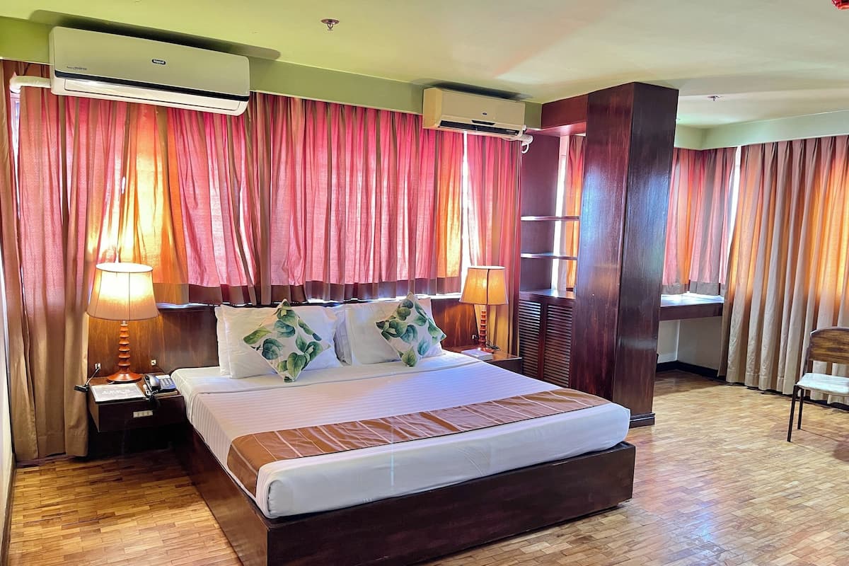 A hotel room with a large bed, two bedside lamps, an air conditioning unit, and a wooden floor.