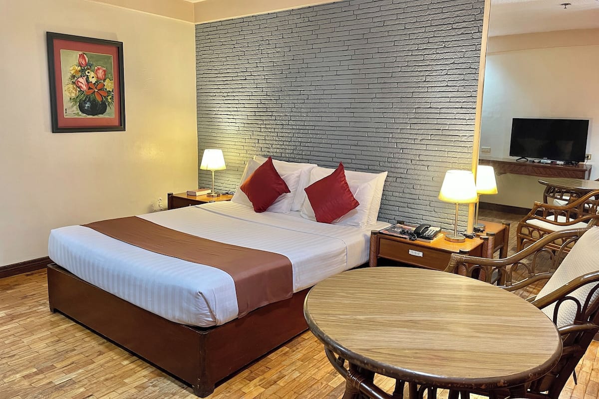 A hotel room with a large bed, two bedside tables with lamps, a round wooden table, wicker chairs, a television, and a brick-patterned headboard.