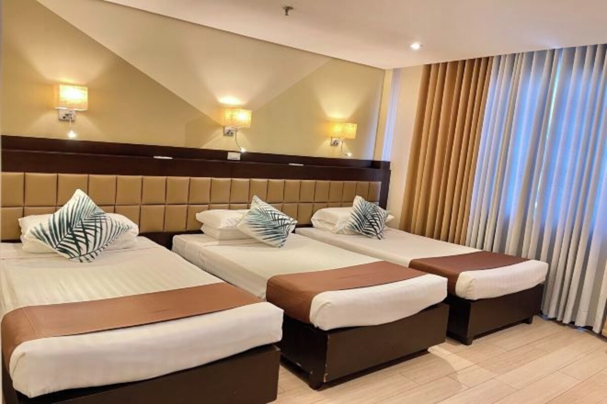 Habitación de hotel con tres camas, cada una con cabecero marrón y ropa de cama blanca. Hay dos luces fijadas a la pared y un espejo grande sobre las camas.