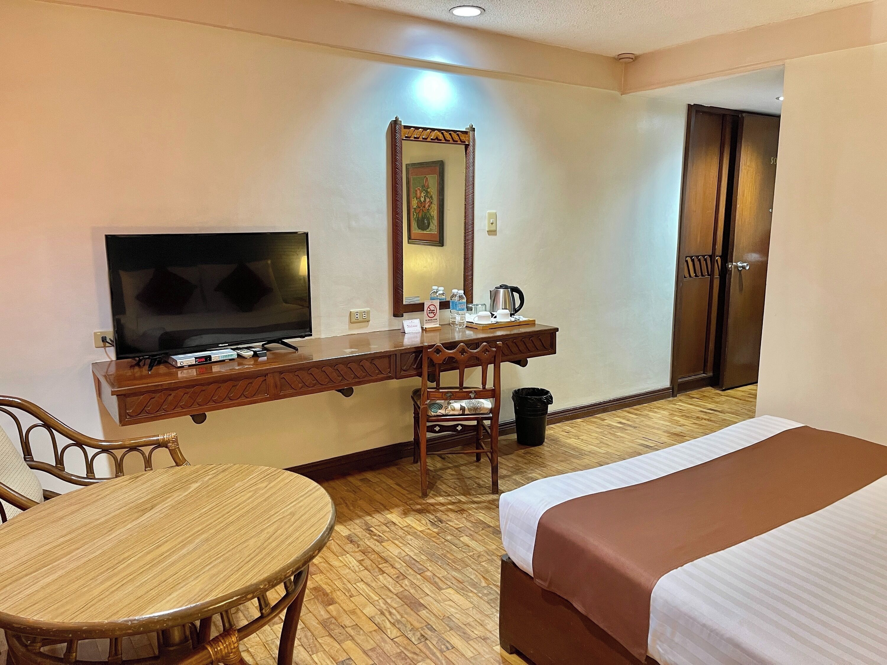Habitación de hotel con cama, escritorio con televisor, silla, mesa, espejo, puerta y cubo de basura.