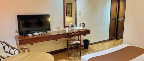 Habitación de hotel con cama, escritorio con televisor, silla, mesa, espejo, puerta y cubo de basura.