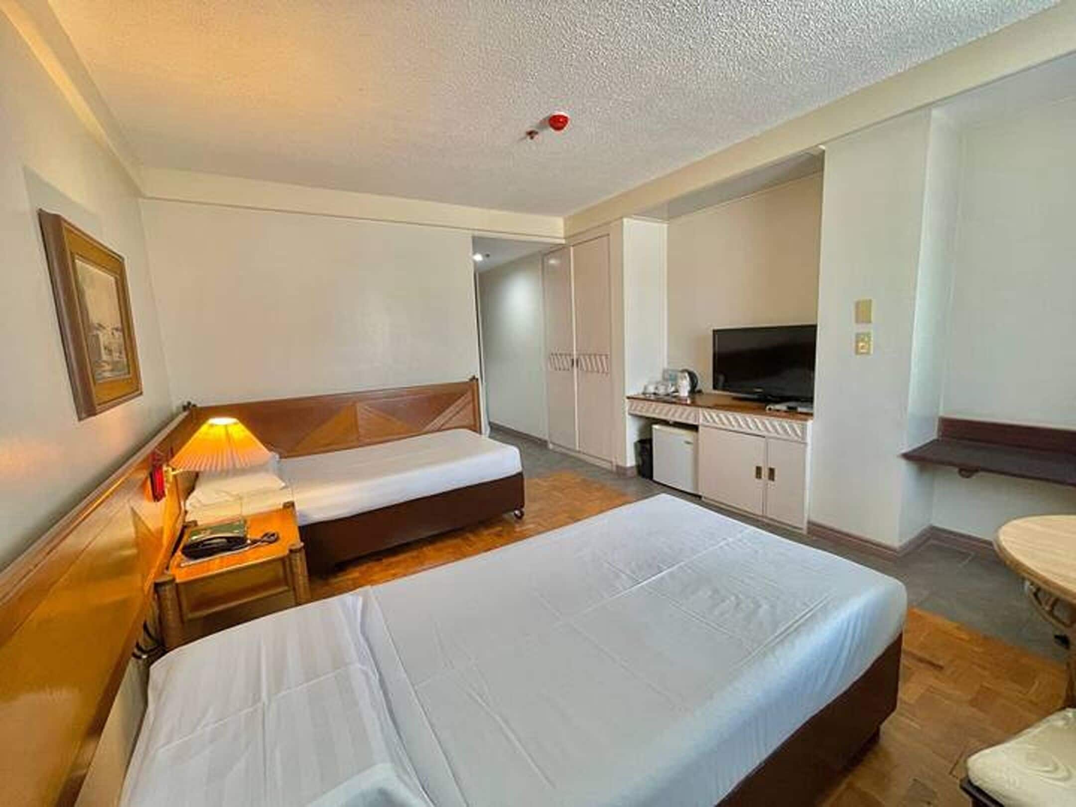 A hotel room with two beds, a desk with a TV, and a small kitchenette.