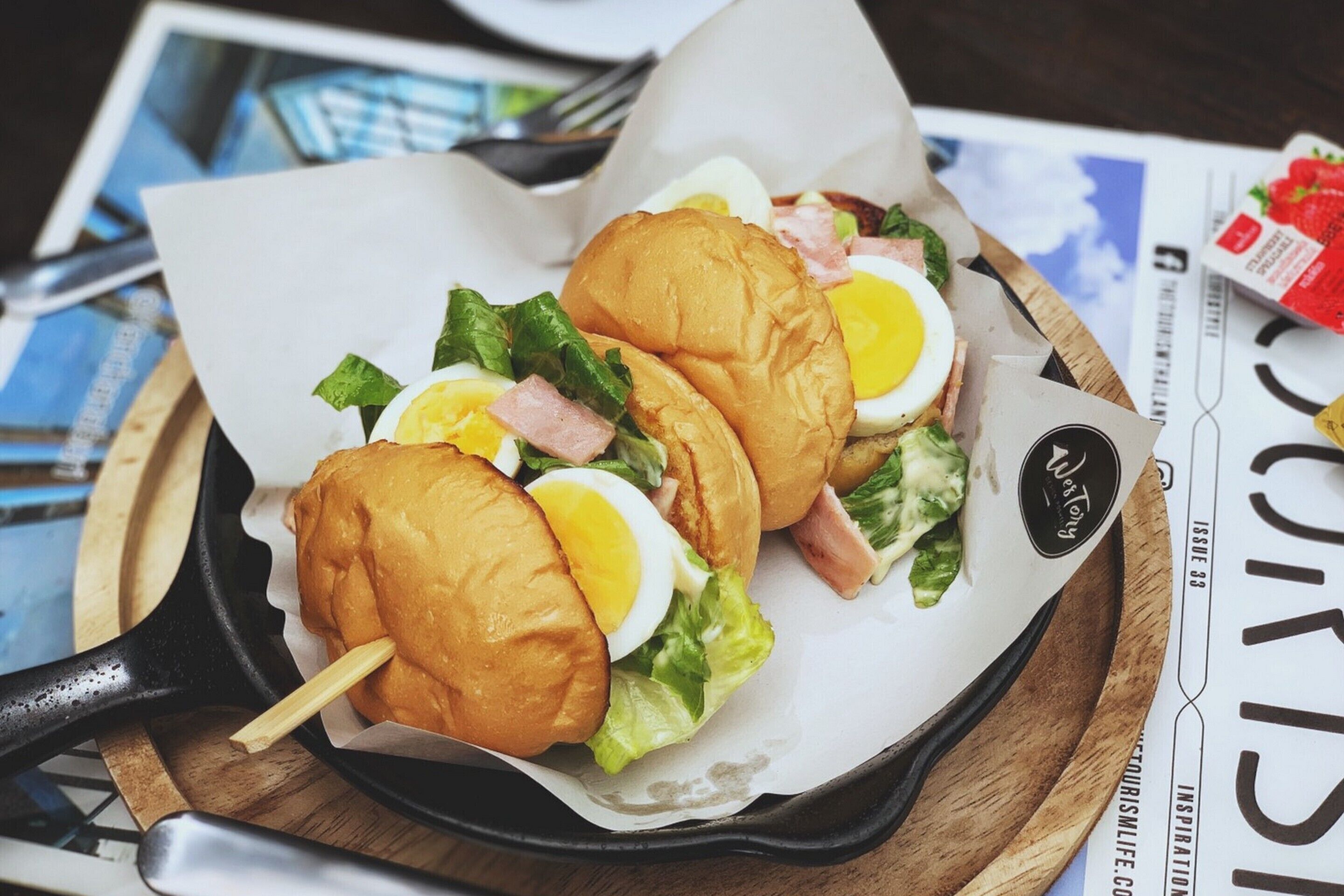 A sandwich with lettuce, egg, and ham on a bun, placed on a wooden board with a black skillet.