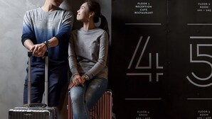 Un hombre de pie junto a una mujer sentada sobre una maleta, con una pared mostrando números y nombres de habitaciones.