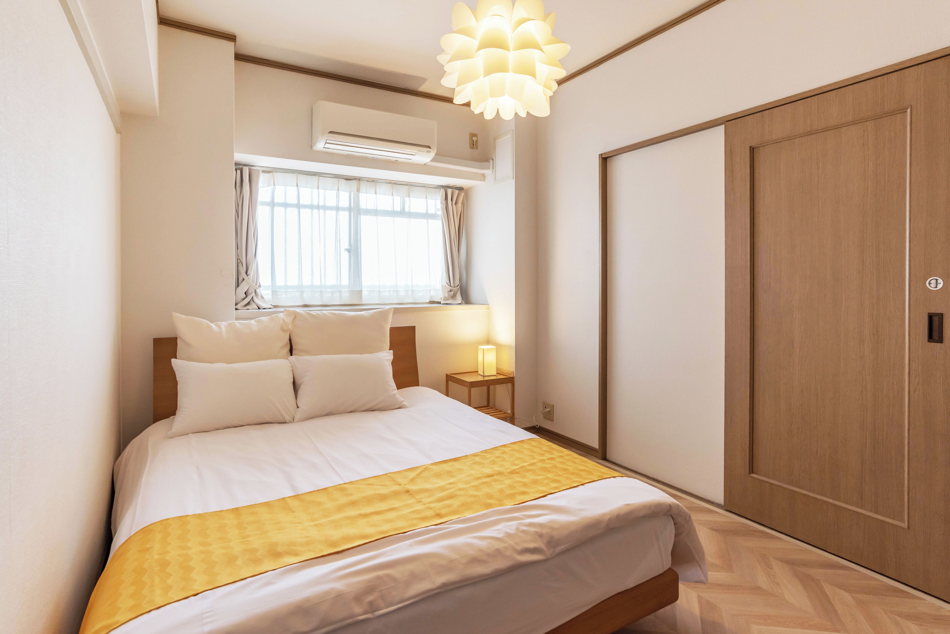 Une chambre d’hôtel avec un grand lit, une table de chevet avec une lampe, un appareil de climatisation, une fenêtre avec des rideaux et une armoire en bois.