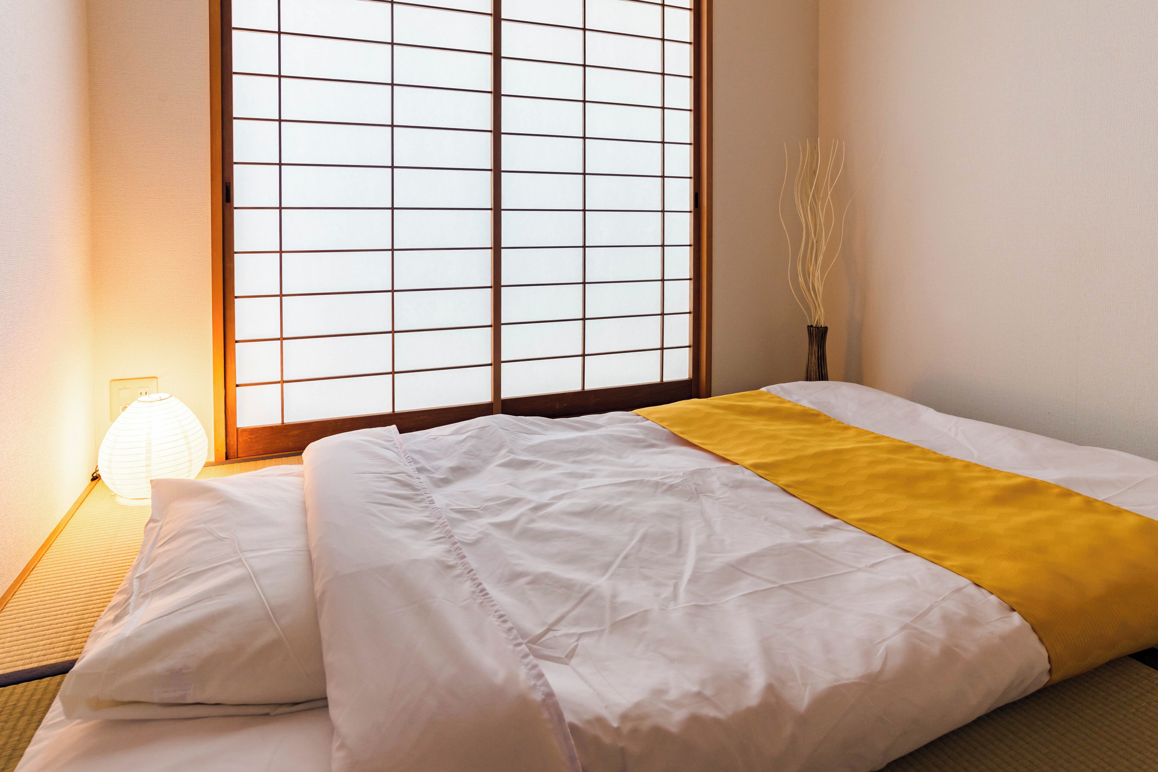 A neatly made bed with white and yellow bedding, a traditional Japanese shoji screen behind it, and a small lamp on the floor beside the bed.