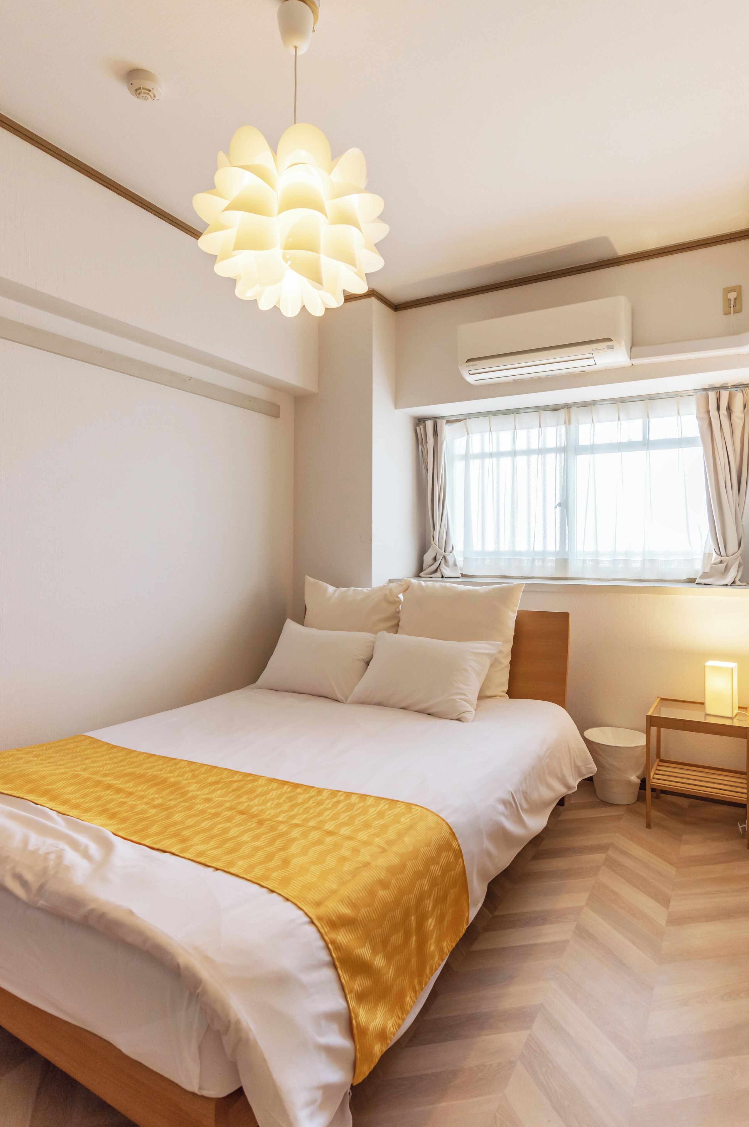 A bedroom with a bed, a bedside table with a lamp, a window with curtains, and an air conditioning unit.