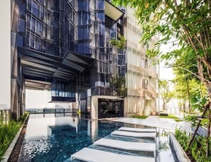 Un edificio moderno con fachada de vidrio reflectante, una piscina en la azotea con escalones y follaje circundante.