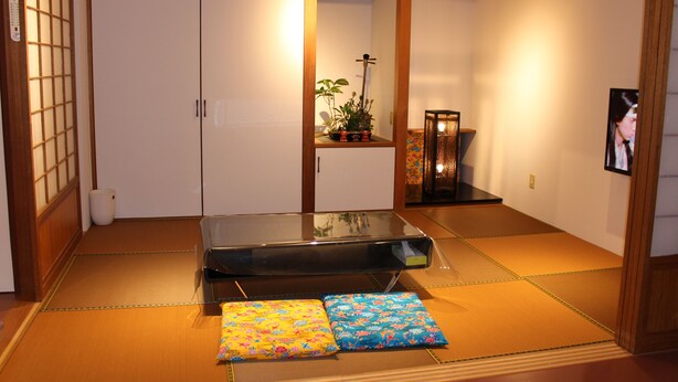 Een traditionele Japanse kamer met tatami vloerbedekking, een lage tafel, kussens, een plank met planten en een televisie.