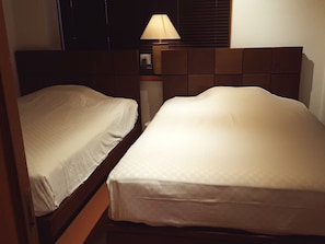 A hotel room with two single beds, a nightstand with a lamp, and a window with blinds.