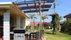 Een terras buiten met een barbecue, een overdekte zithoek en een tuin met palmboompjes en bloemen.
