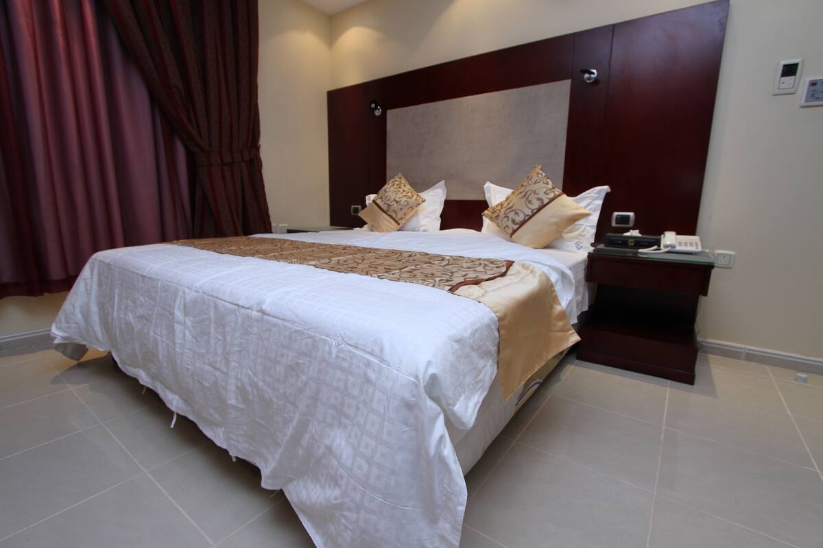 A large bed with a white and beige bedspread, two decorative pillows, and a bedside table with a phone and a small lamp.