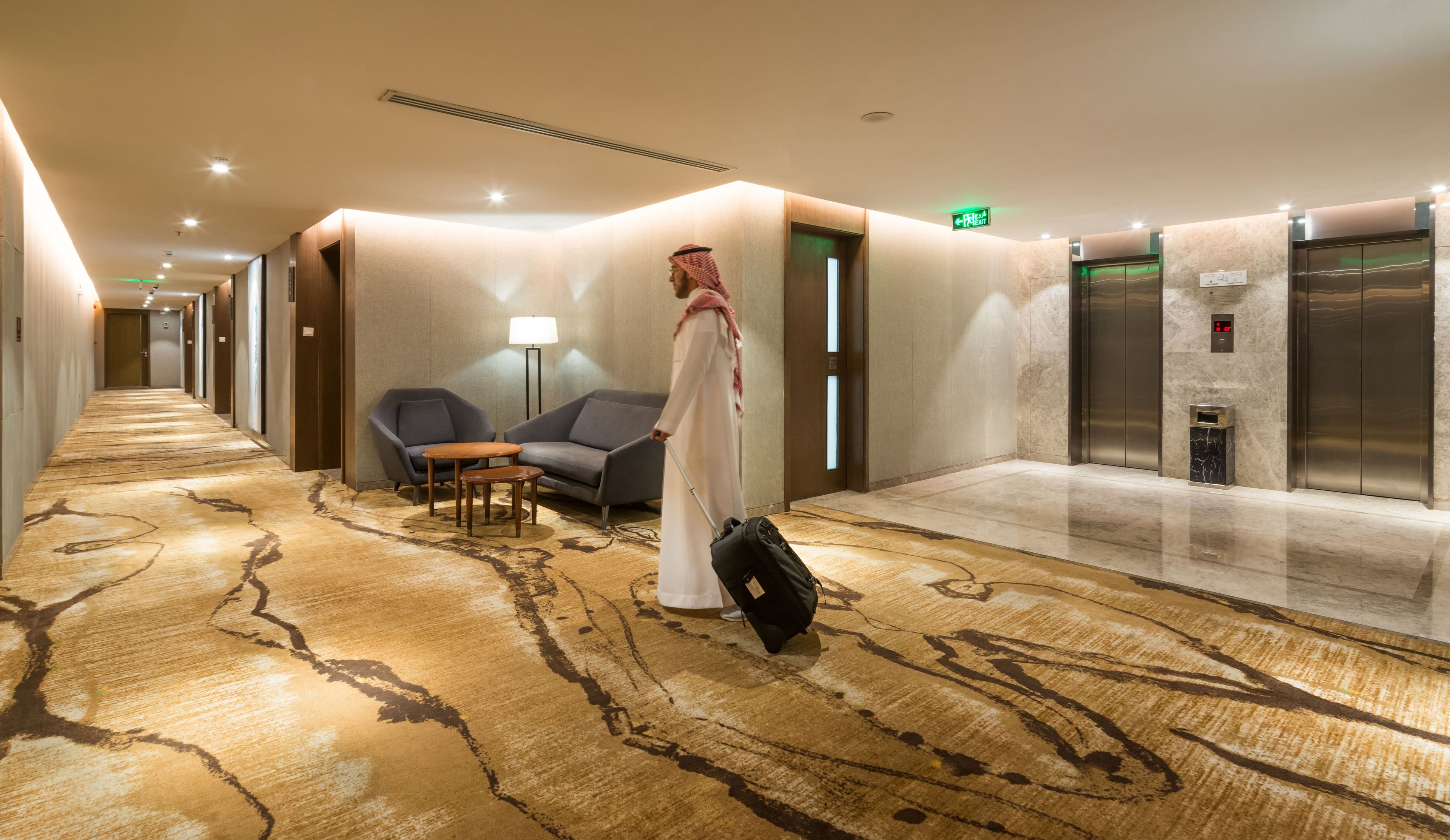 A hotel corridor with a carpeted floor, seating area, and elevators.