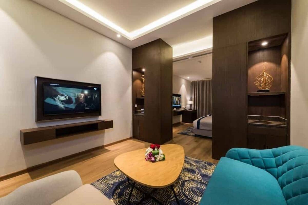 A modern hotel room with a flat-screen TV, a wooden coffee table with flowers, a blue sofa, and a dark wooden accent wall.