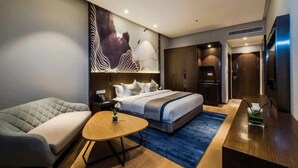 A modern hotel room with a large bed, a sofa, a small round table, and a desk with a chair.