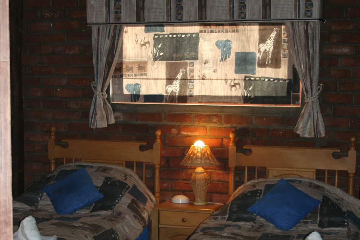 A room with two beds, a brick wall, a window with a view of giraffes, and a lamp on the nightstand.